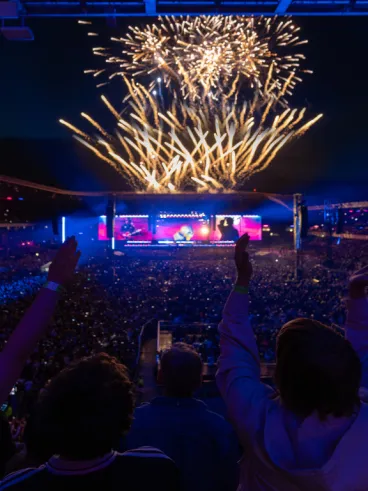 Dark arena with crowd cheering while looking at stage with fireworks overhead