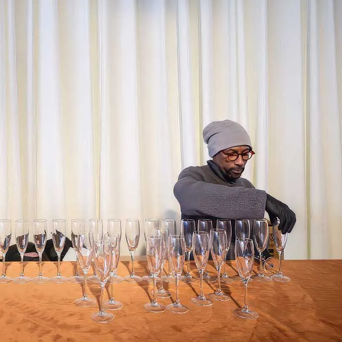 An events staff member in a grey beanie, glasses, and black gloves carefully arranges rows of empty champagne flutes on an orange velvet-draped table, with soft white drapery in the background.