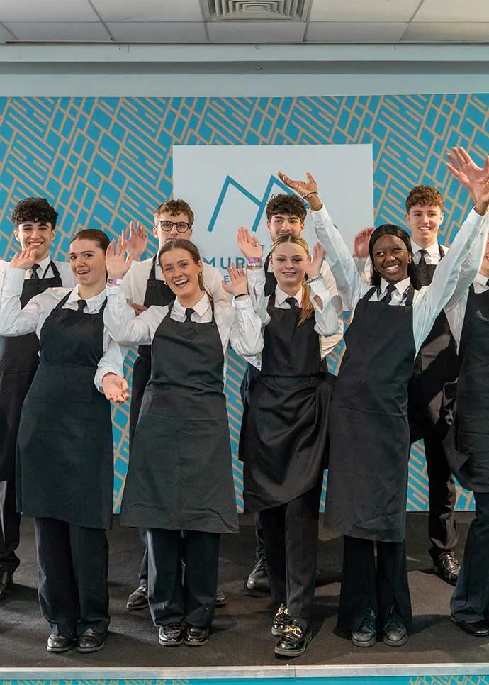 A group of young hospitality staff in white shirts, ties, and black aprons smile and wave at the camera, standing together in front of a branded backdrop with a blue geometric pattern.