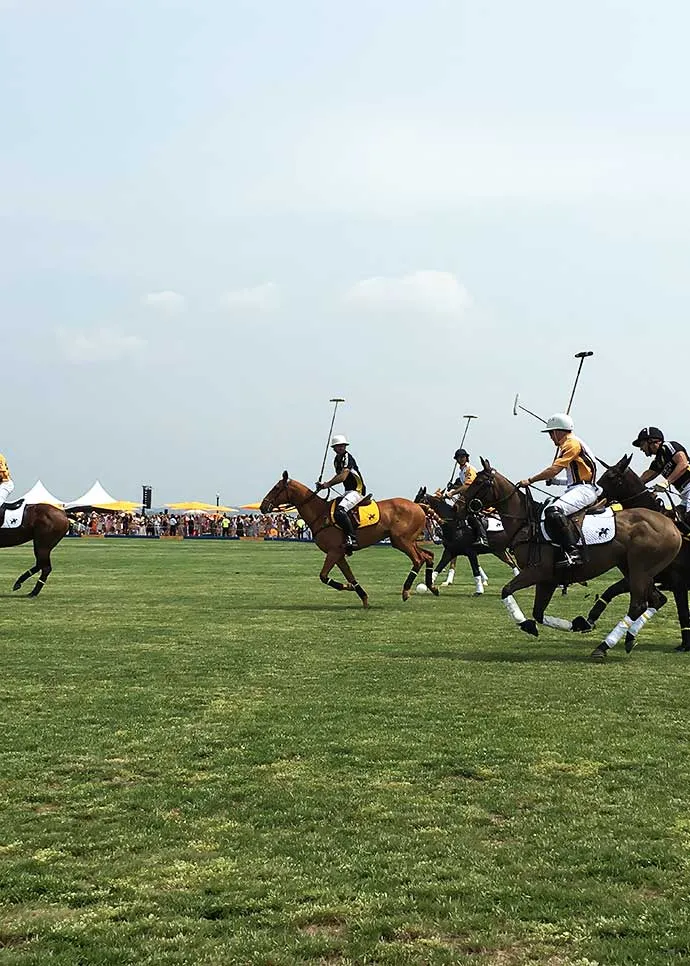 Polo players in yellow and black jerseys ride horses across a grass field during a match, with spectator tents and a crowd visible in the background under a hazy sky.