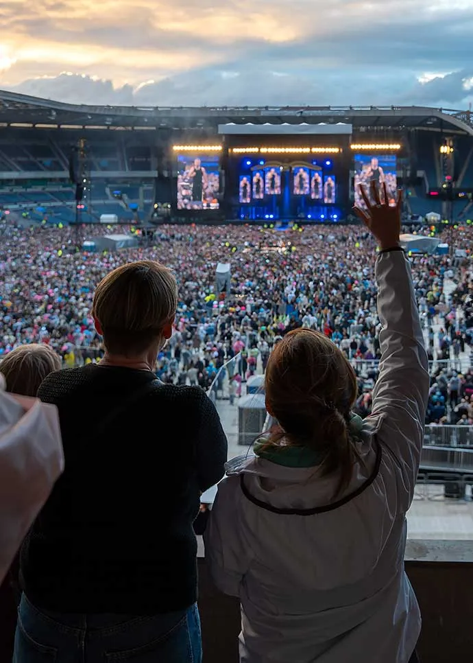 Two concertgoers watch a performance from an elevated vantage point in a packed open-air stadium at dusk, one with her arm raised, as a large stage with video screens lights up in front of a massive crow